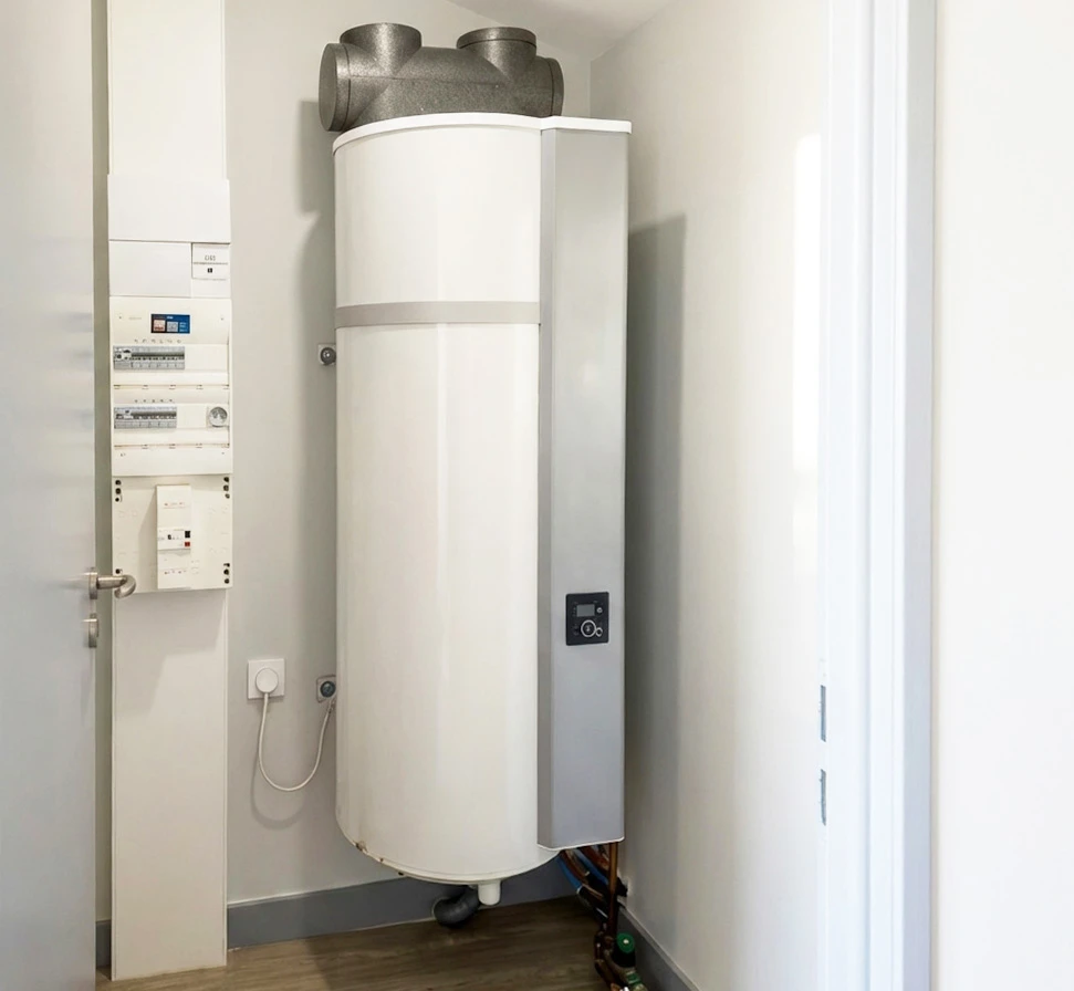 Photographie professionnelle d'une pompe &agrave; chaleur air-air moderne en cours d'installation par un technicien qualifi&eacute; en gilet de chantier, fix&eacute;e &agrave; la fa&ccedil;ade ext&eacute;rieure d'une maison contemporaine. L'unit&eacute; ext&eacute;rieure est en acier inoxydable bross&eacute;, bien visible et clairement identifiable comme &eacute;quipement de chauffage moderne. Le technicien porte des &eacute;quipements de s&eacute;curit&eacute; appropri&eacute;s et manipule l'installation avec professionnalisme. La mise en place claire et professionnelle de ce syst&egrave;me de chauffage moderne repr&eacute;sente la performance &eacute;nerg&eacute;tique et l'&eacute;ligibilit&eacute; aux aides de l'&Eacute;tat.